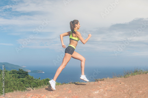 Athletic woman running up the mountain with sky and sea in background. Professional runner doing cardio work-out outdoor in natural landscape