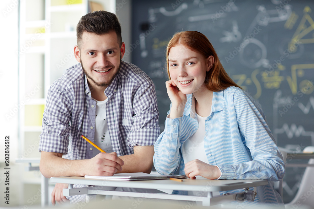 Happy teenage dates sitting by desk in classroom and preparing for next lesson in college Happy teenage dates sitting by desk in classroom and preparing for next lesson in college