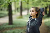 Closeup portrait of content young attractive woman wearing sportswear, raising clasped hands and stretching body in park with green view in background. Front view.
