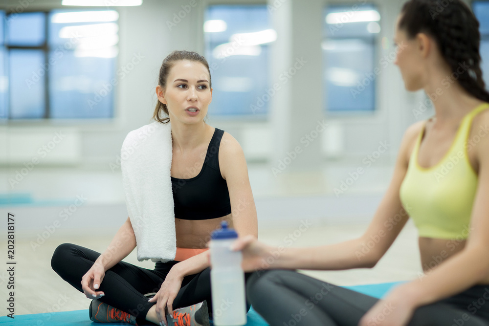 Two friendly girls in activewear sitting on the floor, having talk and refreshment at break between workouts Two friendly girls in activewear sitting on the floor, having talk and refreshment at break between workouts