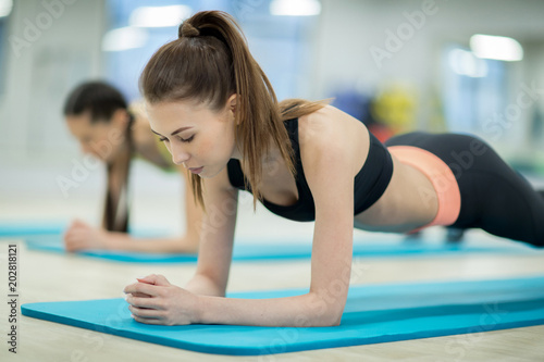 Pretty slim girl in activewear and her friend doing planks on blue mats while training in gym