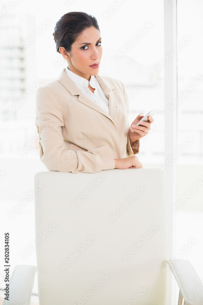Frowning businesswoman standing behind her chair holding her phone glaring at camera Frowning businesswoman standing behind her chair holding her phone glaring at camera