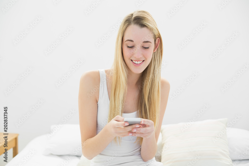 Content blonde woman texting with her smartphone sitting on her bed Content blonde woman texting with her smartphone sitting on her bed