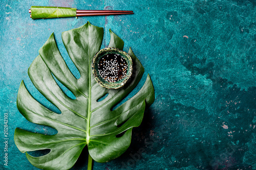 Asian food concept. Chopstick and soysauce soy sauce with white sesame on turquoise background with tropical leaves . Top view