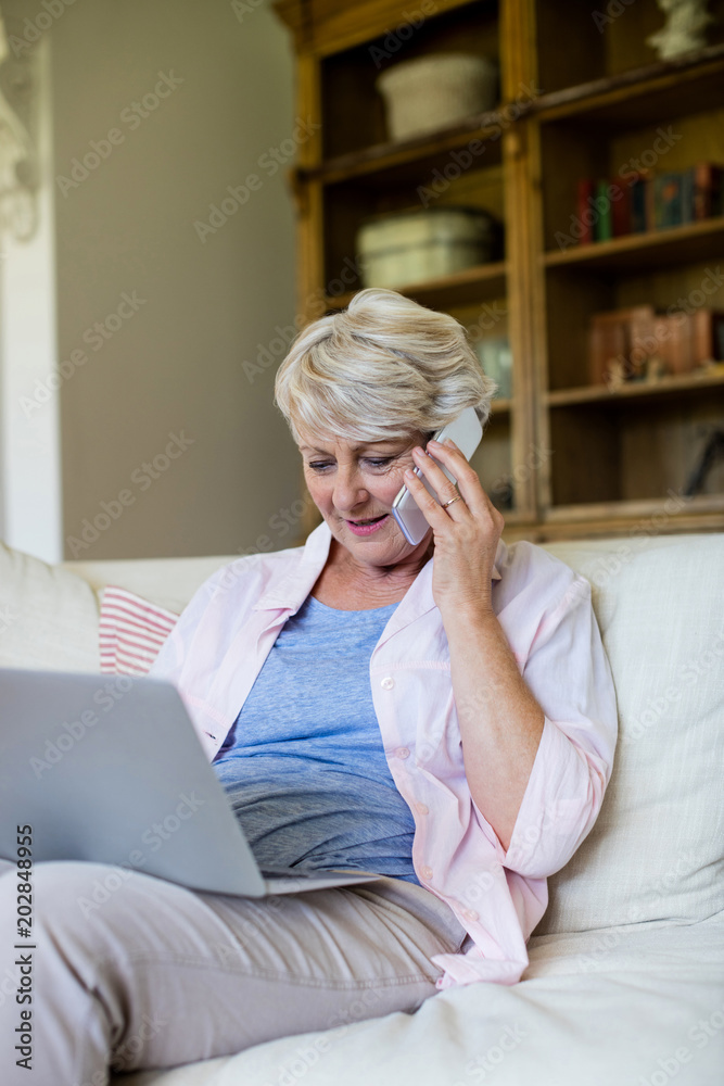 Senior woman talking on mobile phone while using laptop in living room Senior woman talking on mobile phone while using laptop in living room