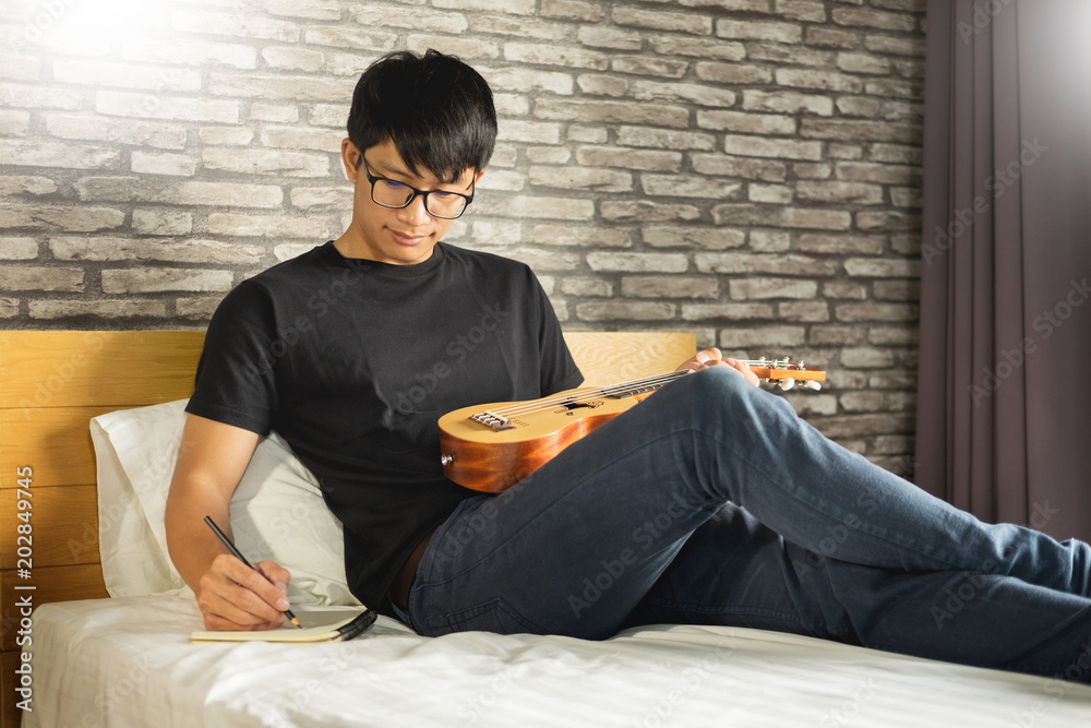 Happy young asian man playing ukulele sitting on bed in bedroom. Happy young asian man playing ukulele sitting on bed in bedroom.