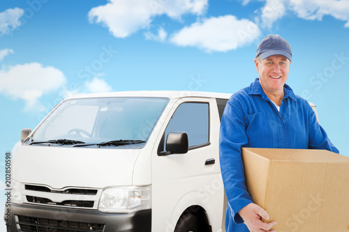 Happy delivery man holding cardboard box against blue sky © vectorfusionart