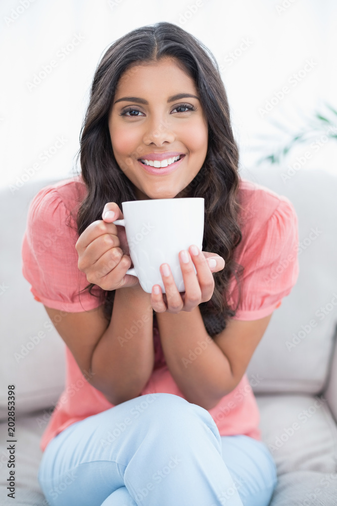 Cheerful cute brunette sitting on couch holding mug Cheerful cute brunette sitting on couch holding mug