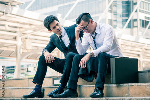 Frustrated Asian young business man sitting outdoor office after he had failed in new project, his colleagues tried to reassure his.