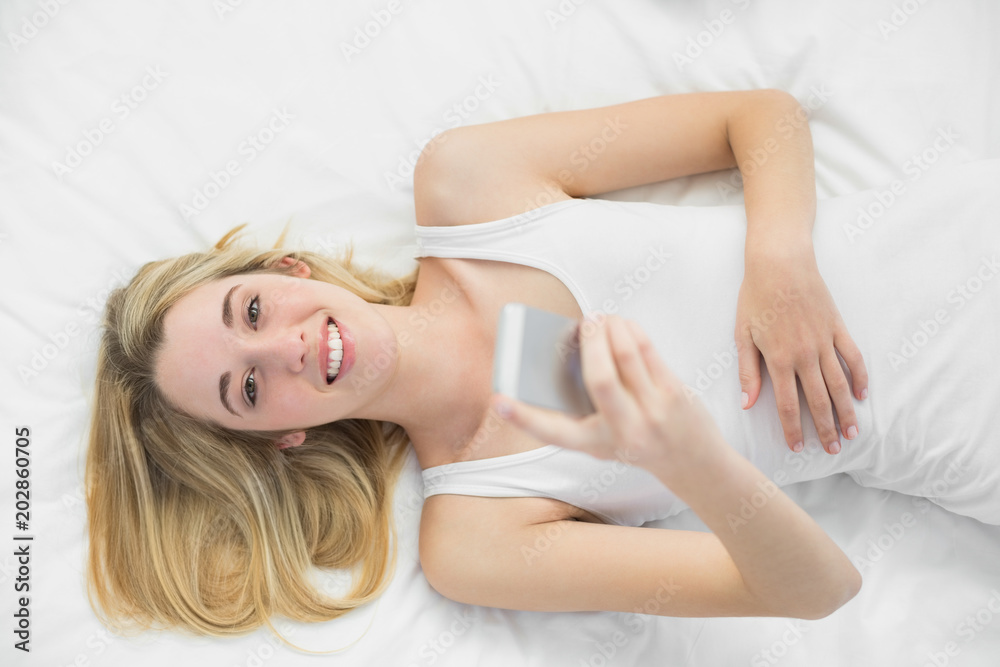 Joyful blonde woman holding her smartphone lying on her bed Joyful blonde woman holding her smartphone lying on her bed
