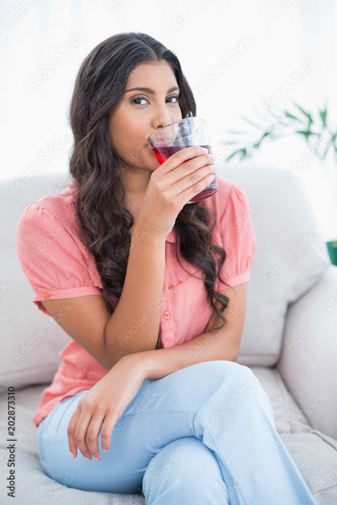 Content cute brunette sitting on couch drinking glass of juice Content cute brunette sitting on couch drinking glass of juice
