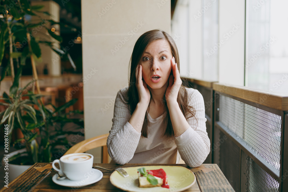 Shocked sad young woman sitting alone near big window in coffee shop at table with cup of cappuccino, cake, relaxing in restaurant during free time. Young female having rest in cafe. Lifestyle concept