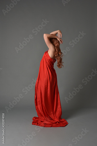 full length portrait of woman wearing red silk gown, on grey studio background.