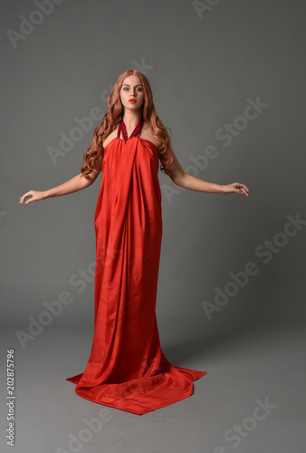 full length portrait of woman wearing red silk gown, on grey studio background.