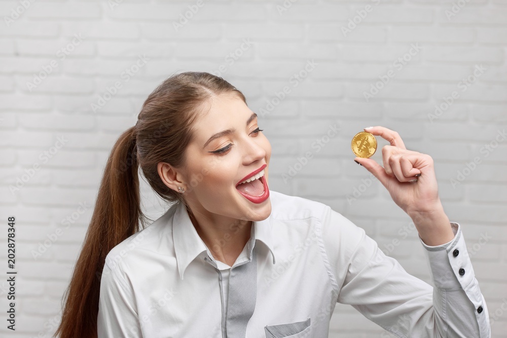 Beautiful positive female finance professional looking with excitement on main cryptocurrency golden bitcoin holding it in hands on blurred background of white brick wall.