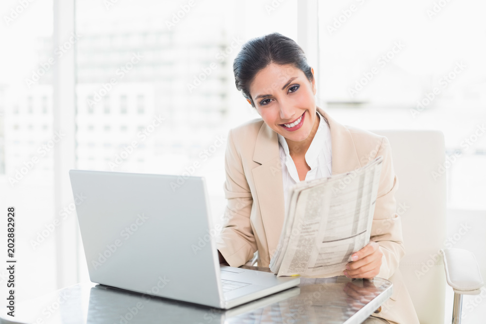 Smiling businesswoman holding newspaper while working on laptop Smiling businesswoman holding newspaper while working on laptop