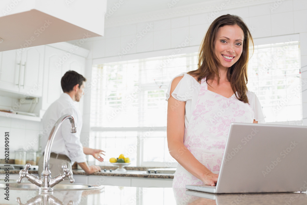 Woman using laptop with man in background in kitchen Woman using laptop with man in background in kitchen