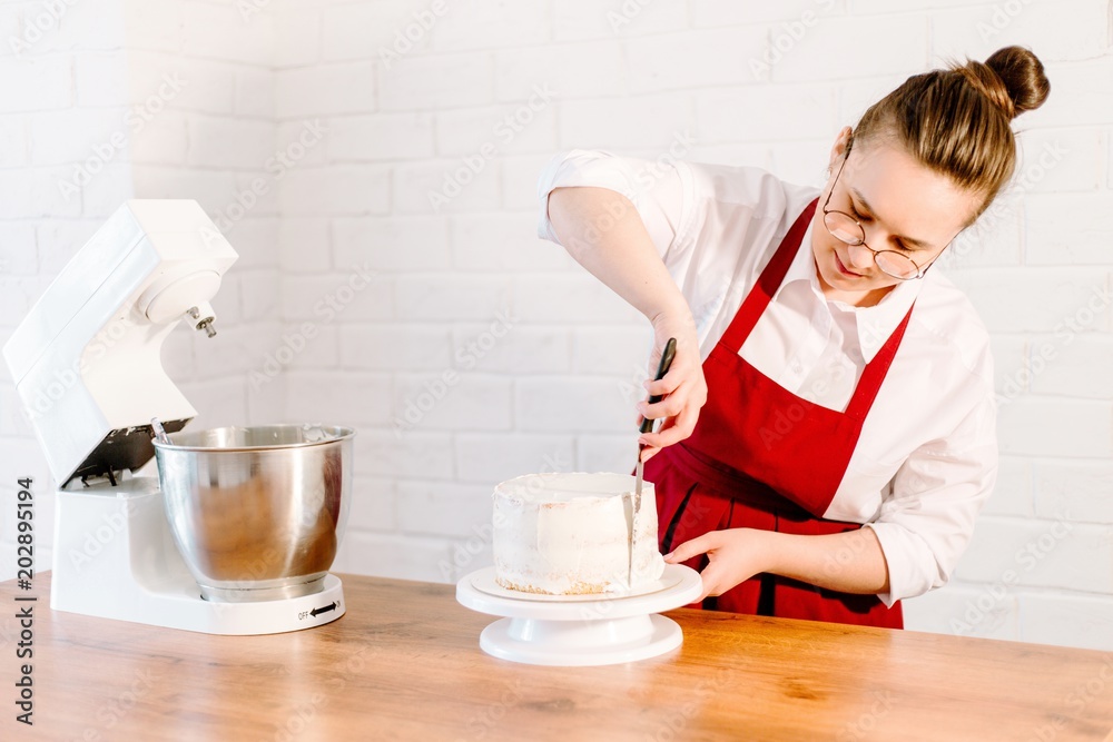 Pastry chef in red apron bakes cake smearing white cream Pastry chef in red apron bakes cake smearing white cream