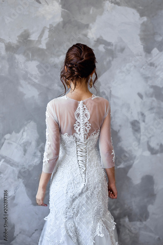 The bride stands with her back against the gray wall. On the bride is a long wedding dress with lace