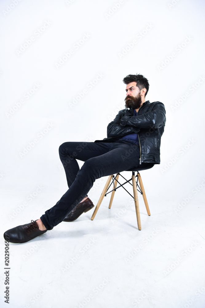 Man with beard and mustache on strict face hold arms crossed. Menswear and fashion concept. Hipster looks serious while sitting on chair in stylish outfit. Macho wears leather jacket, white background