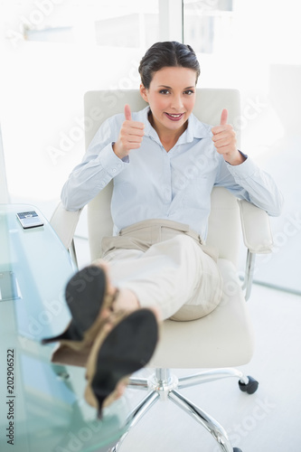 Cheerful stylish brunette businesswoman relaxing with feet up and giving thumbs up