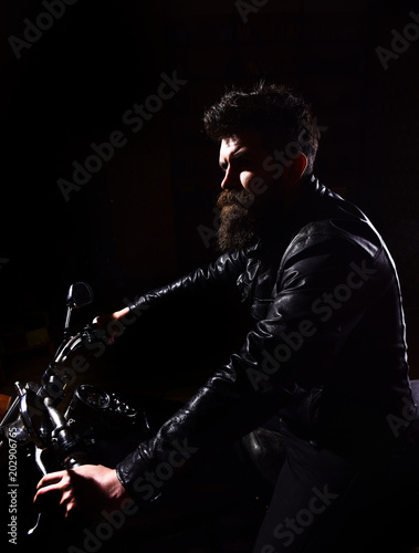 Man with beard, biker in leather jacket sitting on motor bike in darkness, black background. Hipster, brutal biker in leather jacket riding motorcycle at night time, copy space. Night rider concept.