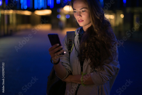 Beautiful young stylish woman walking in the night city streets using modern smartpone