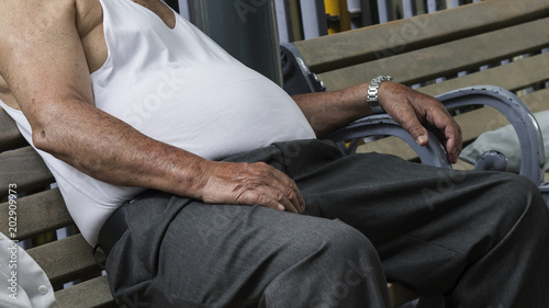 Overweight man in a tank top and trousers slumped on a park bench outdoors. Anonymous figure depicting an unhealthy lifestyle.