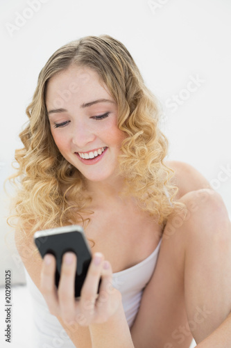 Pretty gleeful blonde sitting on bed holding smartphone