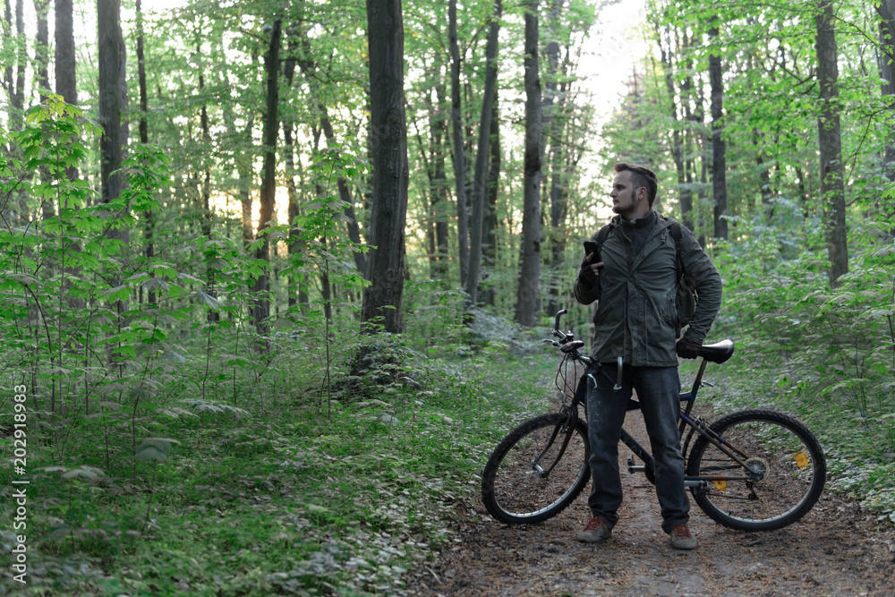Young man with backpack and glasses standing with bicycle on the forest path of the park and looking, active lifestyle concept, sport and jorney, travel surviver concept Young man with backpack and glasses standing with bicycle on the forest path of the park and looking, active lifestyle concept, sport and jorney, travel surviver concept