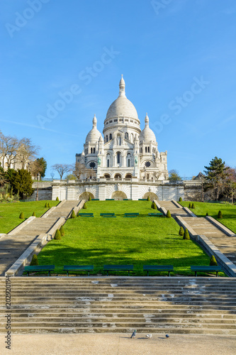 Front view of the bright white basilica of the Sacred Heart of Paris, situated at the top of the Montmartre hill, from the bottom of the stairways in the Louise Michel park by a sunny spring morning. © olrat
