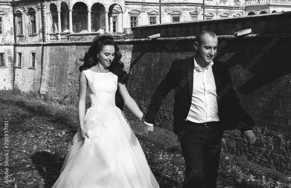 Wedding couple is walking outdoors in sunny autumn day. Beautiful nature and renaissance palace on background. Bride is satin lace dress is holding groom hand. Old castle architecture.