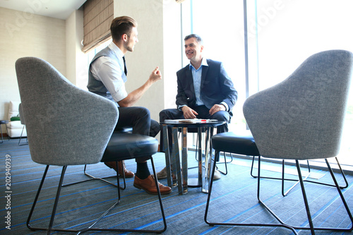 Mature businessman using a digital tablet to discuss information with a younger colleague in a modern business lounge.