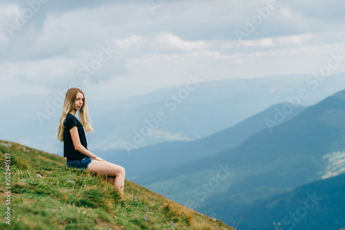 Young girl relaxing and enjoying scenic view on top of mountain. Bliss and happiness. Beautiful summer nature landscape. Lonely young blonde teen traveler dreaming outdoor on vacation. Romantic mood