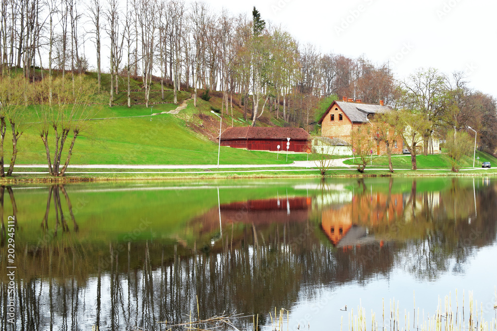 Old red brick house on green hill next to lake with reflection of landscape with trees on water surface.
