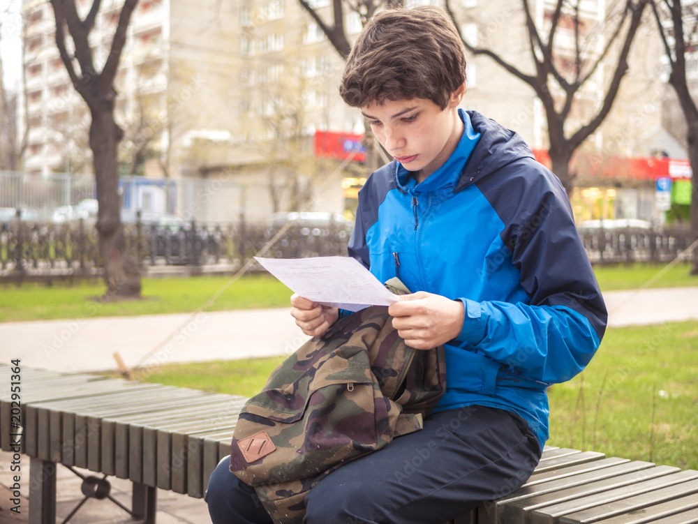 Fototapeta premium portrait of young male teenager sitting on the bench in the street reading paper sheet