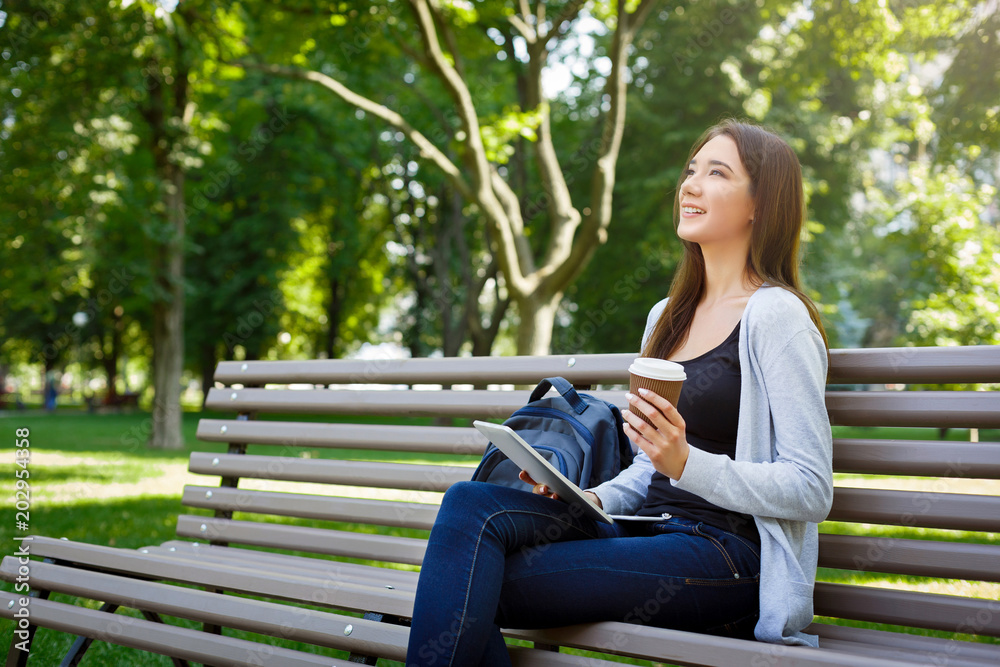 Motivated asian freelancer in the park. Girl on a bench, drinking coffee and working with tablet Motivated asian freelancer in the park. Girl on a bench, drinking coffee and working with tablet