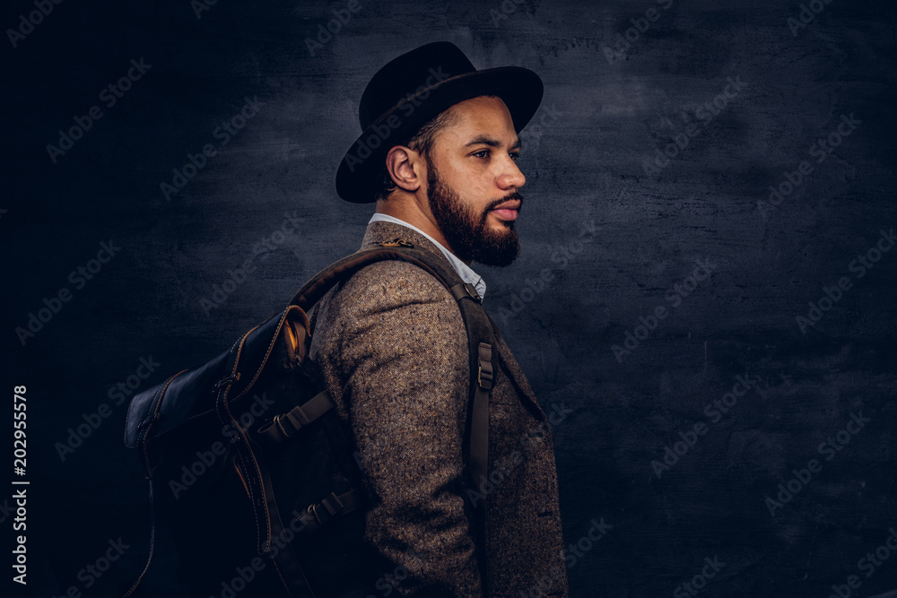 Handsome Afro-American traveler in a brown jacket and hat with the backpack, stands in a studio. Handsome Afro-American traveler in a brown jacket and hat with the backpack, stands in a studio.