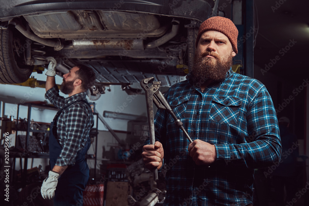 Two bearded brutal mechanics repair a car on a lift in a garage.
