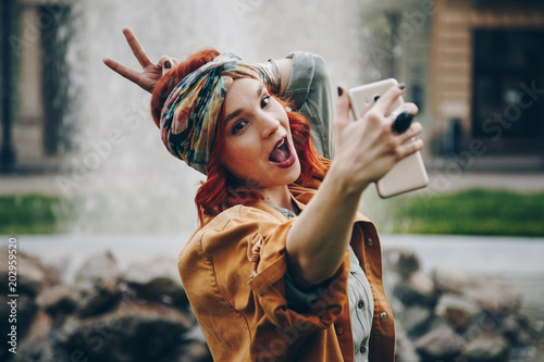 beautiful young woman taking selfie picture outdoors. hippie fashion blogger on vacation, taking self portrait with smartphone. street style, music festival portrait of authentic and fun young girl. photo