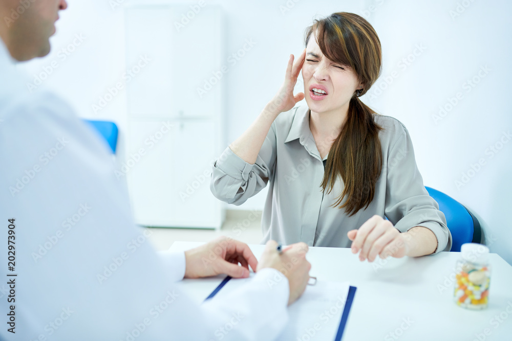 Portrait of beautiful young woman suffering from headache talking to doctor while sitting at desk in clinic, copy space Portrait of beautiful young woman suffering from headache talking to doctor while sitting at desk in clinic, copy space