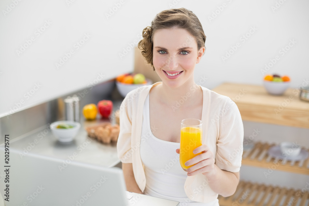 Joyful brunette woman holding a glass of orange juice while sitting in kitchen Joyful brunette woman holding a glass of orange juice while sitting in kitchen