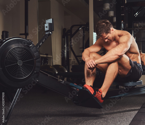 Muscular shirtless athlete resting after a hard workout while sits on the rowing machine in the gym.