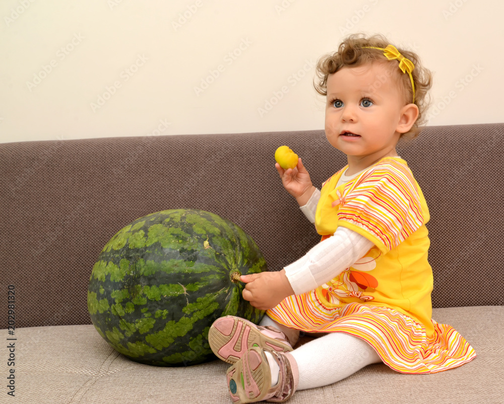 Fototapeta premium The one-year-old girl sits on a sofa and holds with a hand watermelon