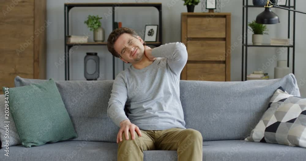 Attractive young caucasian man holding his neck behind as he having headache and feeling pain in the neck and back. At home late at night. Portrait. Indoors Attractive young caucasian man holding his neck behind as he having headache and feeling pain in the neck and back. At home late at night. Portrait. Indoors