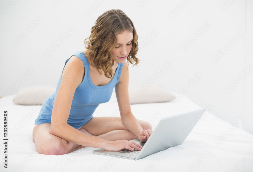 Beautiful serious woman sitting on her bed typing on her notebook Beautiful serious woman sitting on her bed typing on her notebook