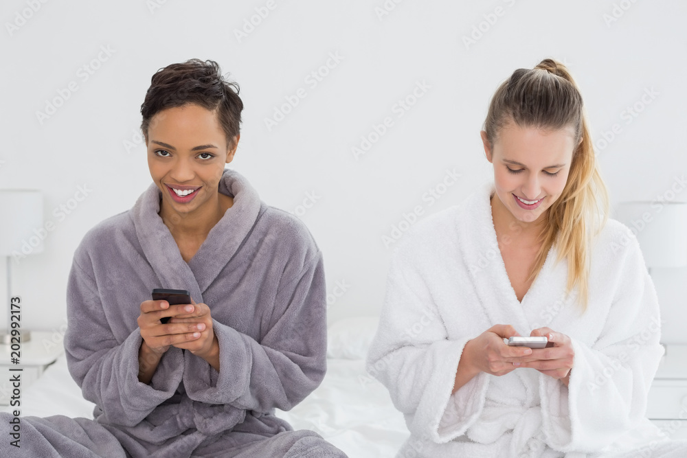 Female friends in bathrobes text messaging on bed Female friends in bathrobes text messaging on bed