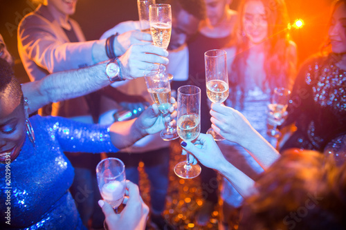 High angle view at multi-ethnic group of laughing young people enjoying dance party in nightclub and drinking champagne, focus on clinking glasses