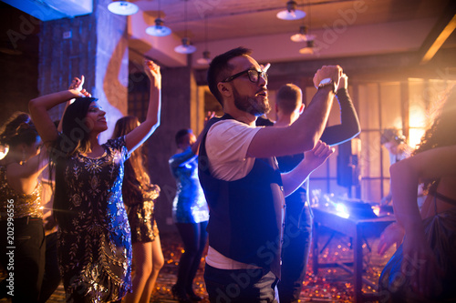 Crowd of trendy people dancing in nightclub with golden confetti flying around, focus on Asian man dancing in center enjoying party