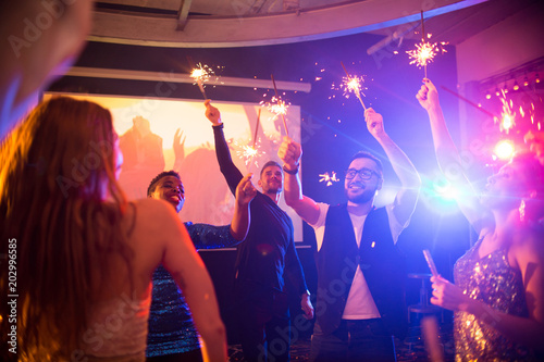 Group of trendy young people celebrating holiday in nightclub burning flaming sparklers on dance floor  copy space